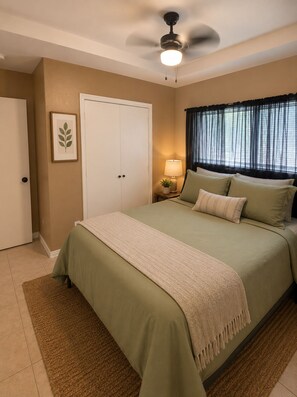 Une chambre à coucher avec un lit, un ventilateur de plafond, une fenêtre avec des rideaux, une porte et un tableau encadré au mur.