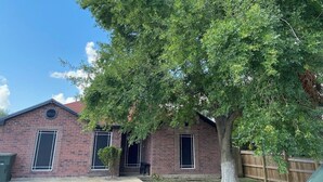 A brick building with a large tree in front, a wooden fence, and a clear blue sky. - Perfect 3-bedroom house with AC in welcoming Harlingen (Harlingen)