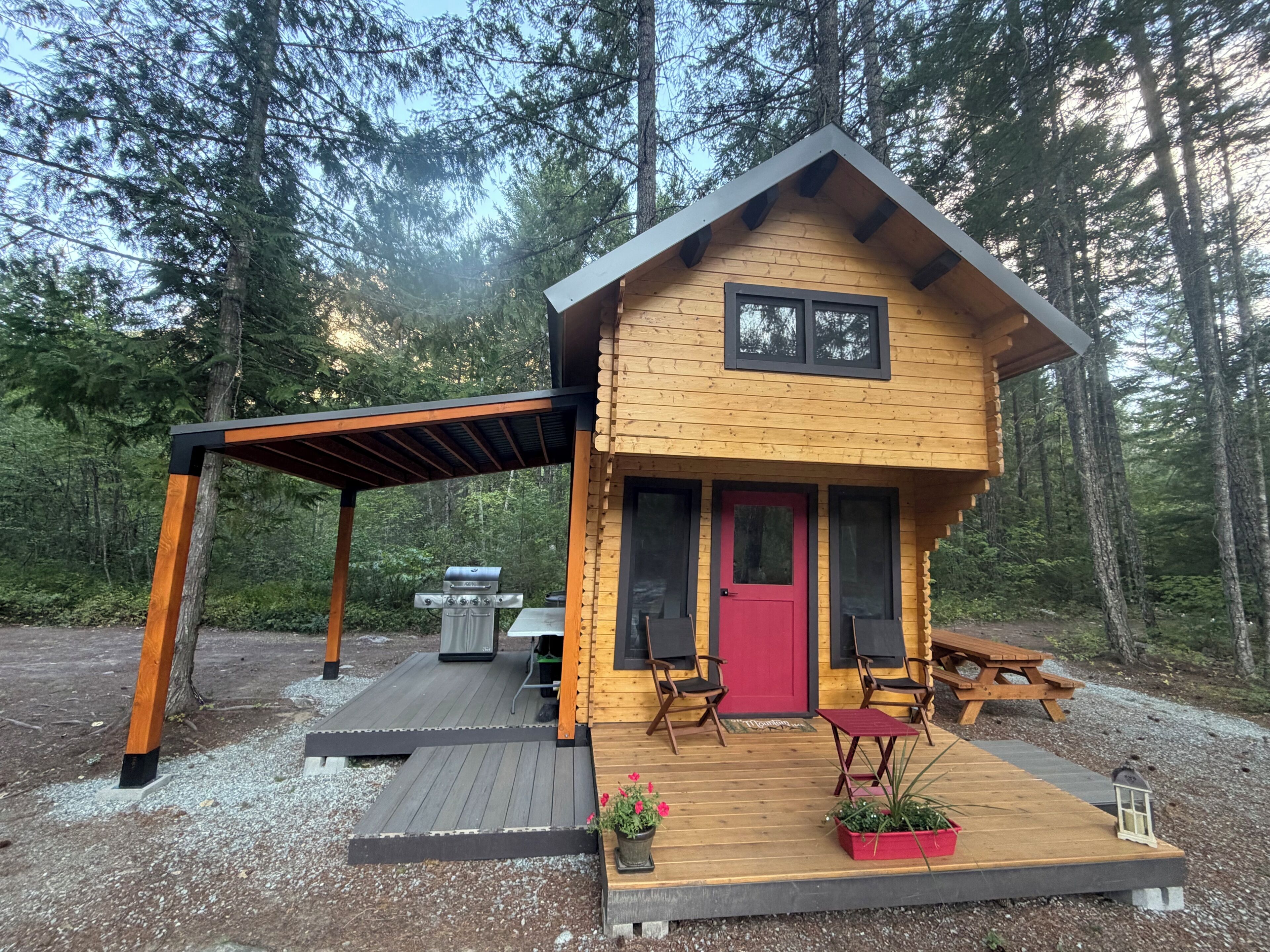 A wooden cabin with a red door, two chairs, a table, and a grill outside.