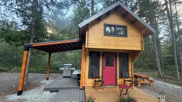 A wooden cabin with a red door, two chairs, a table, and a grill outside.