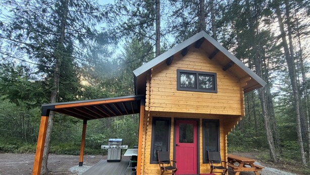 A wooden cabin with a red door, two chairs, a table, and a grill outside.