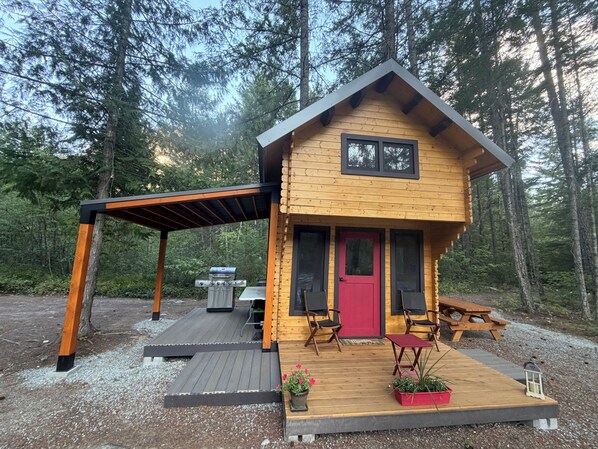 A wooden cabin with a red door, two chairs, a table, and a grill outside.