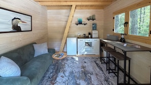 A compact cabin with a wooden interior, a kitchenette, a sofa, and a large window offering a view of the forest.