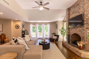 A living room with a brick fireplace, a ceiling fan, and a large TV mounted on the wall. - Hidden Falls Retreat: Private, Blowing Rock, Hot Tub (Lenoir)