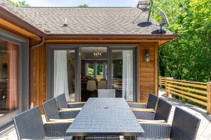A covered outdoor dining area with a table and chairs, a wooden deck, and a glass door leading to an interior space.