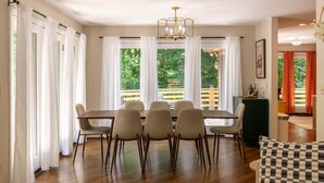 A dining area with a table and chairs, large windows with curtains, a chandelier, and a view of greenery outside. - Hidden Falls | Private, Near Blowing Rock , Hot tub (Lenoir)