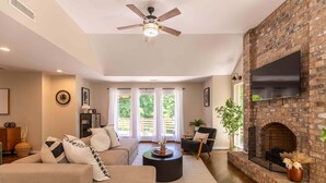 A living room with a brick fireplace, a ceiling fan, and a large TV mounted on the wall. - Hidden Falls | Private, Near Blowing Rock , Hot tub (Lenoir)