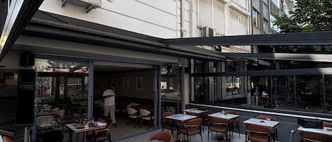 Uma área externa com mesas e cadeiras, uma seção coberta e um edifício com várias janelas e aparelhos de ar condicionado.