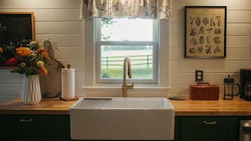 Una cocina con un lavabo blanco, una ventana con un adornado floral, una ilustración enmarcada de un huerto y un jarrón de flores en la encimera.