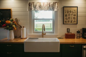 Una cocina con un lavabo blanco, una ventana con un adornado floral, una ilustración enmarcada de un huerto y un jarrón de flores en la encimera.