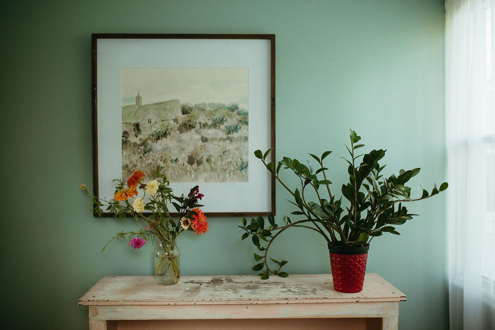 Un quadro incorniciato, un vaso con fiori e una pianta in vaso su un tavolo, di fronte a una parete verde.