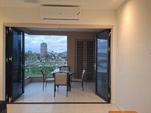 Un balcón con mesa y sillas, un equipo de aire acondicionado y vistas a un paisaje urbano con edificios y zonas verdes.