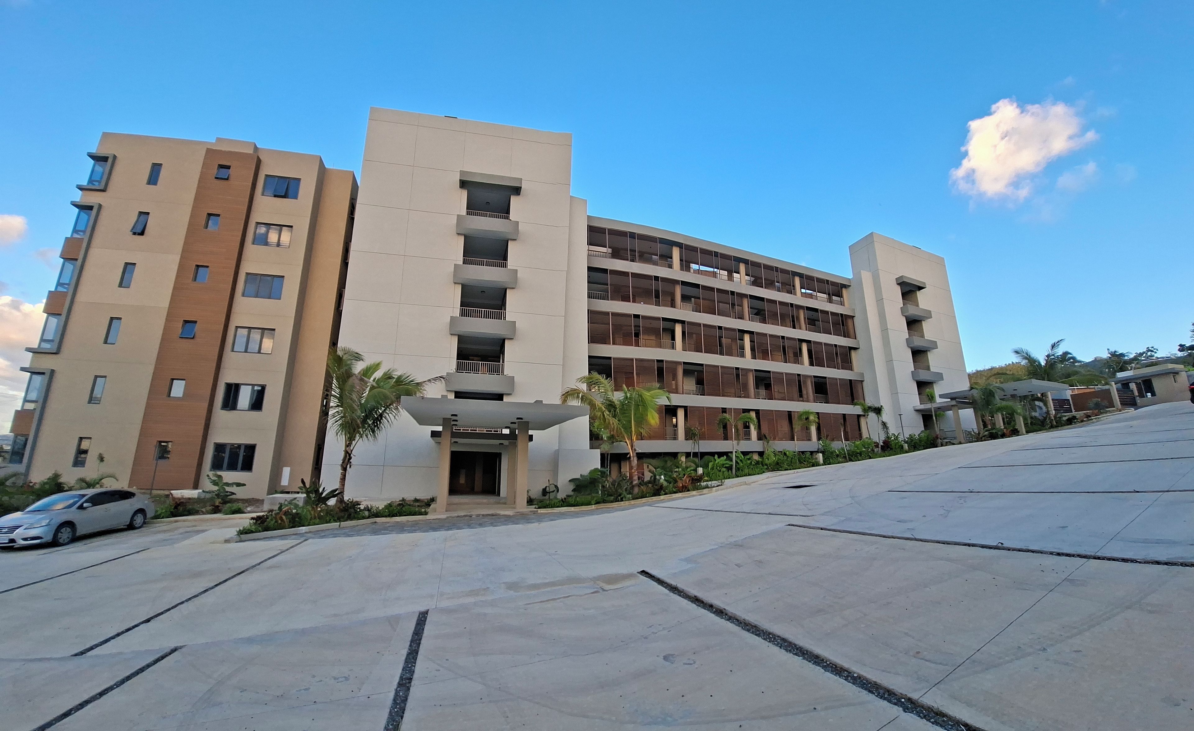A multi-story residential building with balconies, a parking area, and a clear sky.