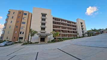 A multi-story residential building with balconies, a parking area, and a clear sky.