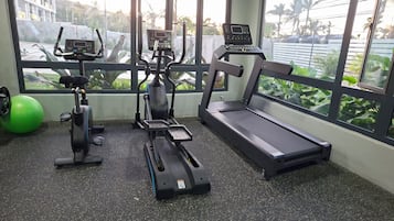 A gym with cardio equipment, a green exercise ball, and a window with a view of palm trees.