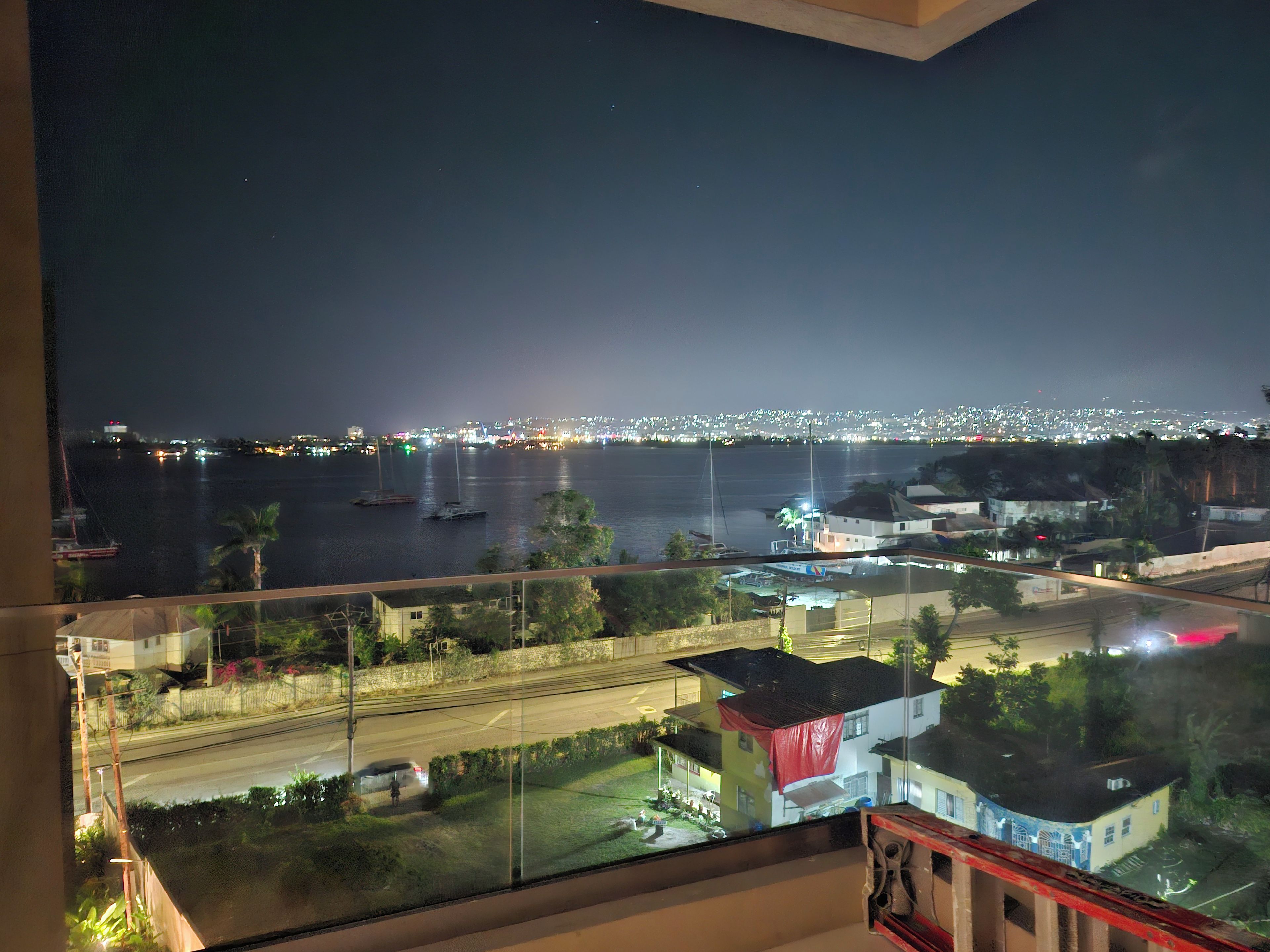 A nighttime view from a balcony overlooking a body of water with boats, a city skyline in the distance, and a road with vehicles.