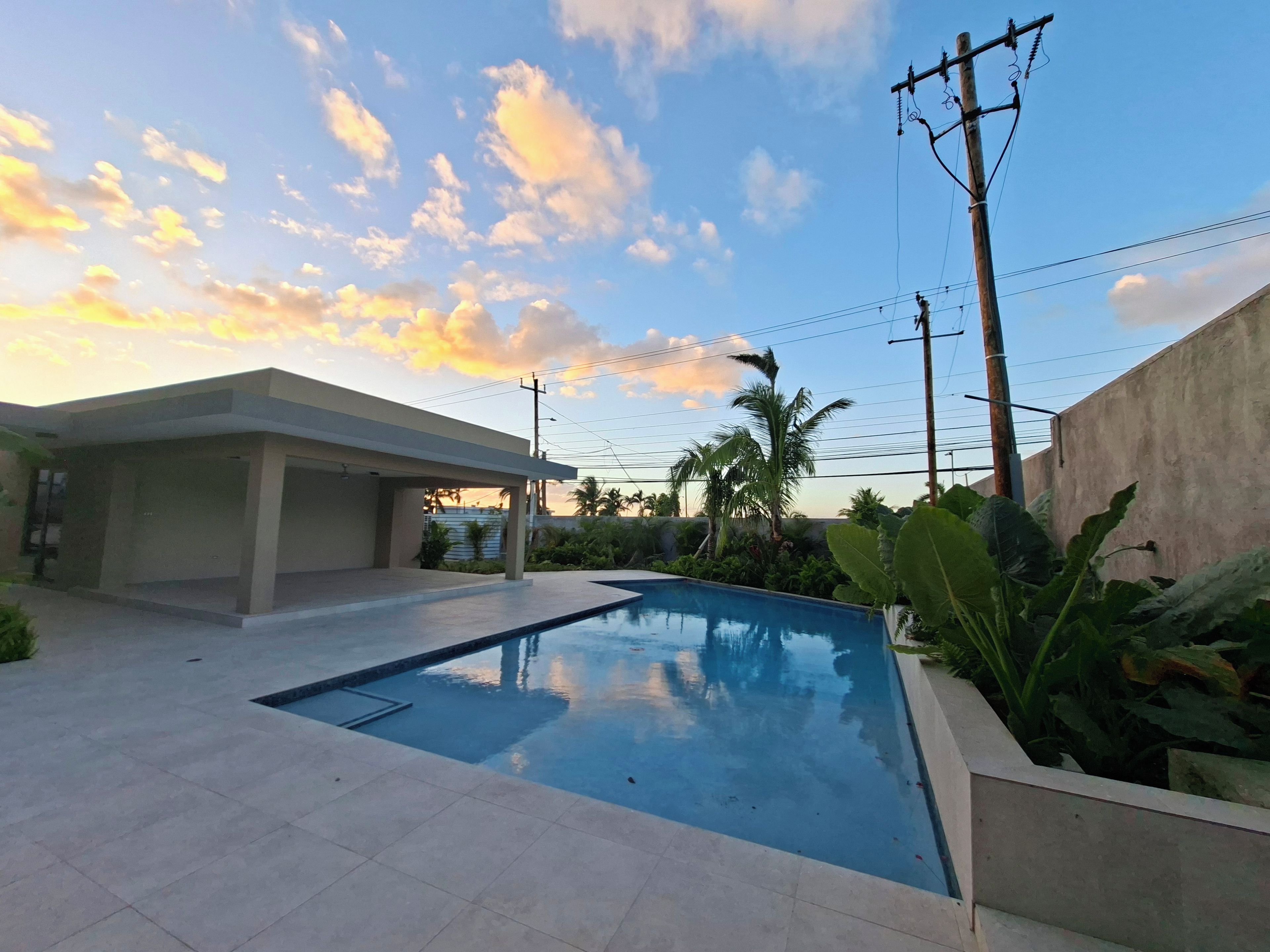 A swimming pool with a tiled deck, surrounded by greenery and a building with a flat roof.