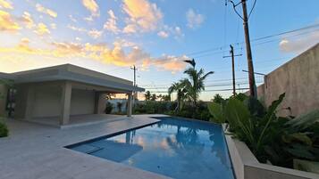 A swimming pool with a tiled deck, surrounded by greenery and a building with a flat roof.