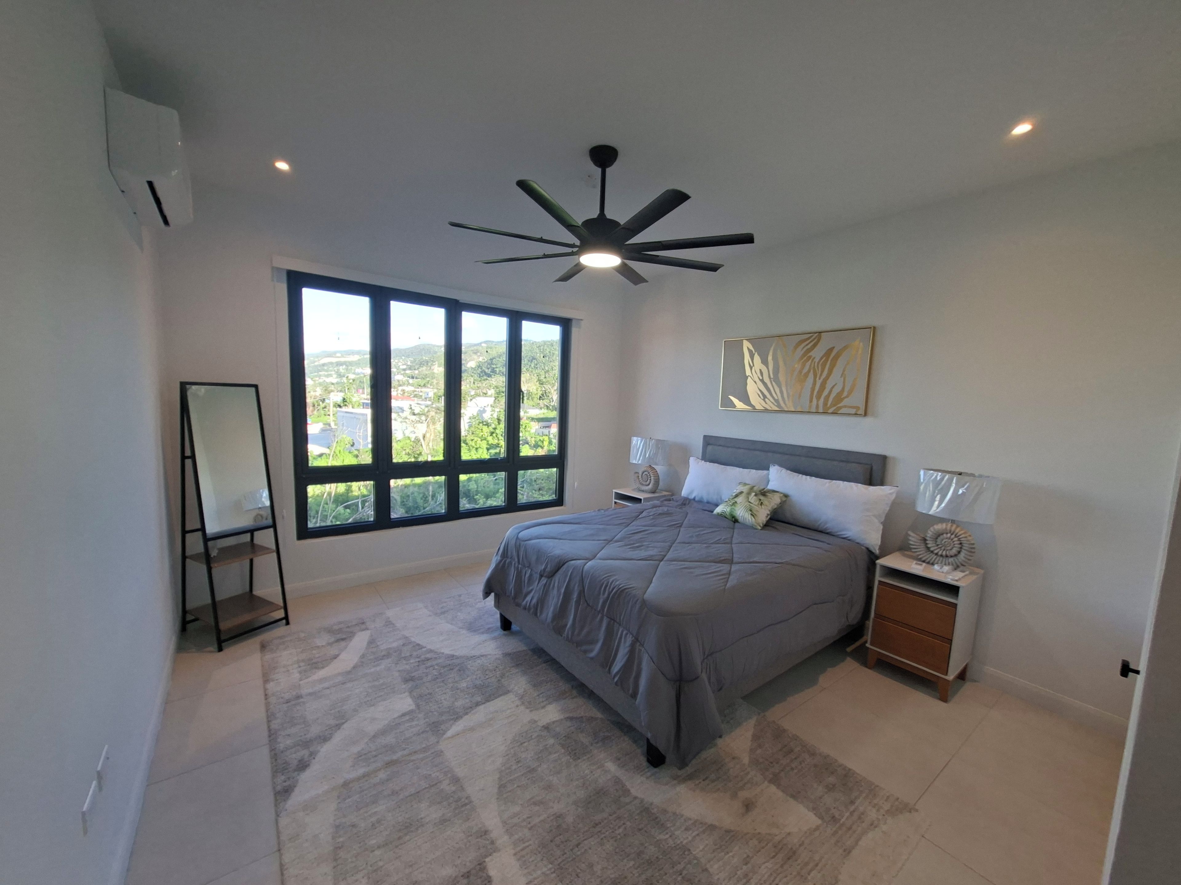 A bedroom with a large window, a ceiling fan, a bed with a grey bedspread, two bedside tables with lamps, and a decorative wall piece.