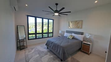 A bedroom with a large window, a ceiling fan, a bed with a grey bedspread, two bedside tables with lamps, and a decorative wall piece.