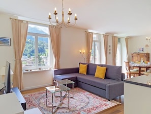 Ein Wohnzimmer mit einer Couch, einem Glastisch, einer Pendelleuchte und Blick auf eine gebirgige Landschaft durch große Fenster.