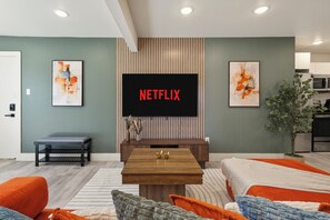 Ein modernes Wohnzimmer mit einem großen Flachbildfernseher, einem Holztisch und einer Couch mit orangefarbenen Kissen.
