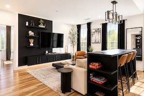 Una sala de estar moderna con un mueble de entretenimiento negro, un televisor de pantalla plana, un sofá, una mesa de centro y un área de comedor con taburetes de bar.