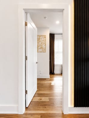 Un pasillo con suelo de madera, paredes blancas, una puerta, una lámpara en el techo y una ventana con cortinas.