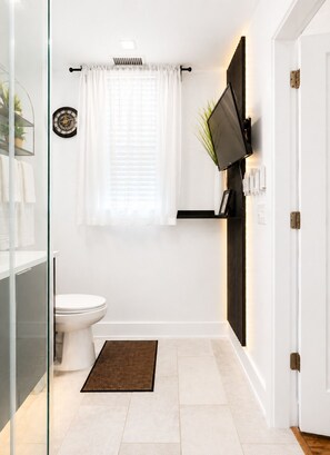 A modern bathroom with a toilet, a shower area, a glass door, a white tiled floor, and a mounted television. - Midnight Luxe Suite with Walkable Areas and Free Parking (Atlanta)