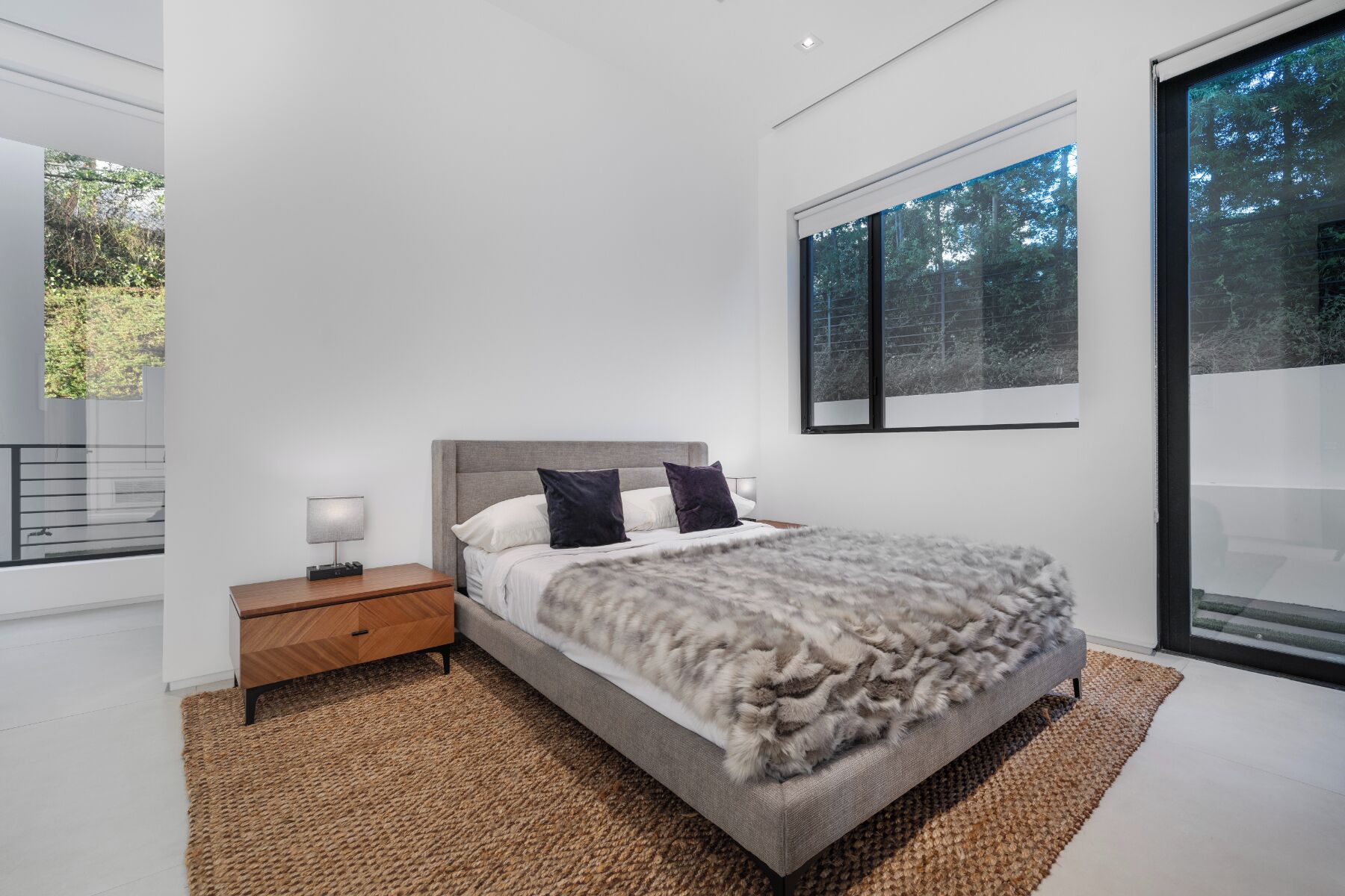 A modern bedroom with a large bed, a nightstand, a window with a view of trees, and a balcony.