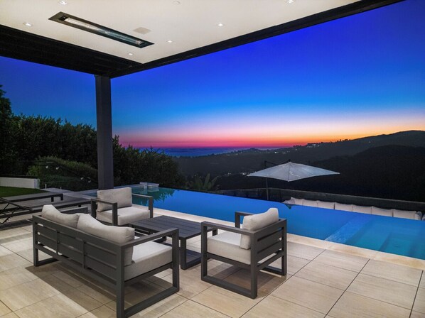 Une terrasse extérieure moderne, dotée d’une piscine, d’un espace salon et offrant une vue sur un coucher de soleil se déclinant sur un paysage vallonné.