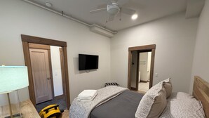 A bedroom with a bed, a TV, a ceiling fan, and a door leading to a bathroom.