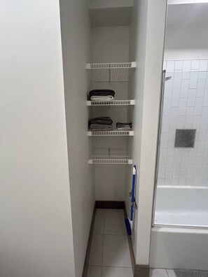 Ein schmaler Flur mit weißen Fliesenböden und -wänden, der zu einem Badezimmer mit Dusche und Toilette führt. Drei weiße Regale mit gefalteten Handtüchern sind vorhanden.