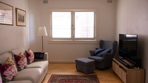 A living room with a sofa, ottoman, armchair, TV, and a window with blinds. - Double Bay Art Deco Apartment with Harbour Vistas (Double Bay)