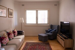 Ein Wohnzimmer mit Sofa, Hocker, Sessel, Fernseher und einem Fenster mit Jalousien.