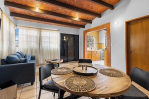 A dining area with a round wooden table, black chairs, and woven placemats. There is a sofa and a window with curtains in the background. - Chic little house in San Miguel (San Miguel de Allende)