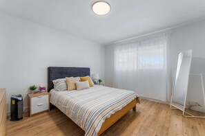 Een slaapkamer met een bed, nachtkastjes, een nachtlampje, een staande lamp, een raam met dunne gordijnen en een whiteboard.
