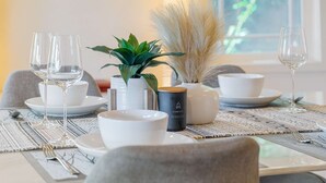 Una mesa de comedor dispuesta con platos, fuentes y vasos blancos, junto con una planta en maceta y objetos decorativos.