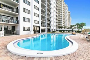 Piscina com placa de 'NÃO MERGULHAR', rodeada por um edifício branco com varandas e um céu azul límpido.