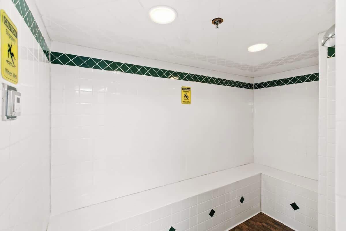 Un baño con paredes de azulejos blancos y un borde con diseño de diamantes verdes. Hay dos carteles: uno advierte sobre pisos resbaladizos y otro indica piso mojado.