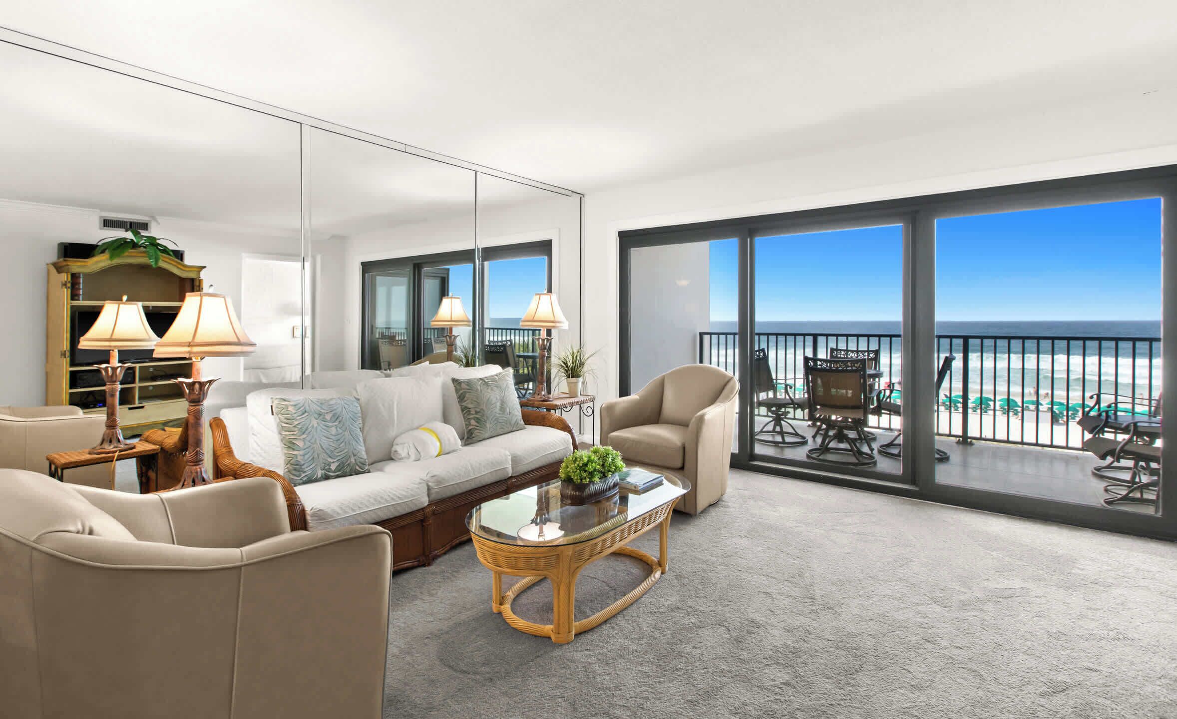 A modern living room with a sofa, chairs, a glass coffee table, and a view of the beach through large windows.