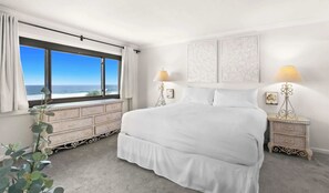 Quarto com uma cama grande, duas mesas de cabeceira, cômoda, janela com vista para o oceano e arte nas paredes.
