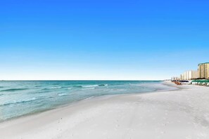 Uma praia com céu claro, areal arenoso e uma fileira de edifícios ao fundo.
