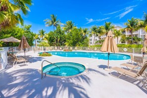 A swimming pool with lounge chairs and umbrellas, surrounded by palm trees and a clear blue sky.
