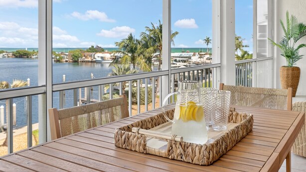 Een houten tafel met een gevlochten dienblad, een waterkan en glazen op een balkon met uitzicht op een jachthaven en de oceaan.