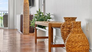 Un suelo de madera, una lámpara de pie tejida, un banco tejido y una planta en maceta en una habitación con paredes blancas.