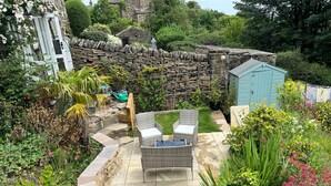 Un mur de pierres, un petit abri de jardin et une terrasse avec des meubles en osier.