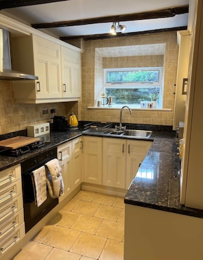 A kitchen with white cabinets, a granite countertop, a built-in stove, a sink, and a window with a view of greenery. - 17th Century Cottage, Log Burner & Hidden Garden (Laycock)