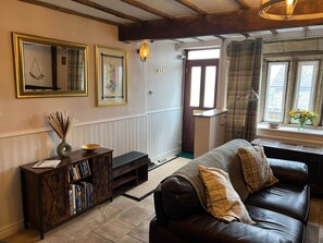 A living room with a dark leather sofa, a wooden console table with books, a vase with dried plants, and a window with curtains. - 17th Century Cottage, Log Burner & Hidden Garden (Laycock)