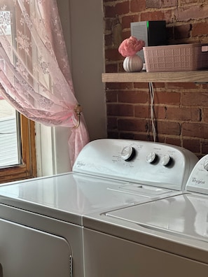 A white Whirlpool washer in a room with a brick wall, a window with pink curtains, and a wooden shelf with a basket and a vase.